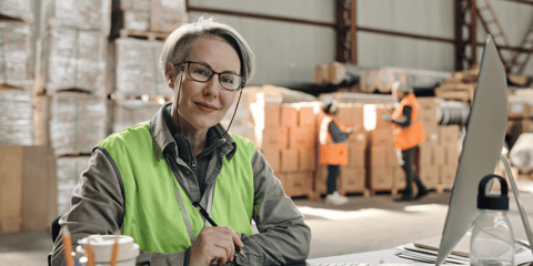 Female logistics professional sitting at a desk in a warehouse, smiling at the camera